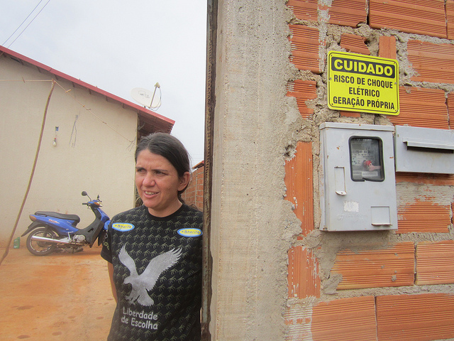Patricia Soares de Oliveira, who was the first to receive solar panels as a test in 2017, stands in front of her house and next to an electric meter that reads "danger of electric shock". Her power bill in this social housing complex on the outskirts of Palmeiras de Goiás in central Brazil has fallen to one-fifth of what she previously paid. Credit: Mario Osava/IPS