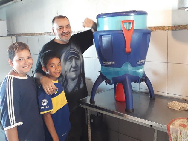 Julio Esquivel and two children in the La Casita de La Virgen soup kitchen in Villa La Cava stand next to the filter that removes 99.9 percent of bacteria, viruses and parasites, with a capacity of up to 12 liters per hour. The purifier became the starting point for raising awareness in this shantytown on the outskirts of the Argentine capital about access to water as a human right. Credit: Daniel Gutman/IPS