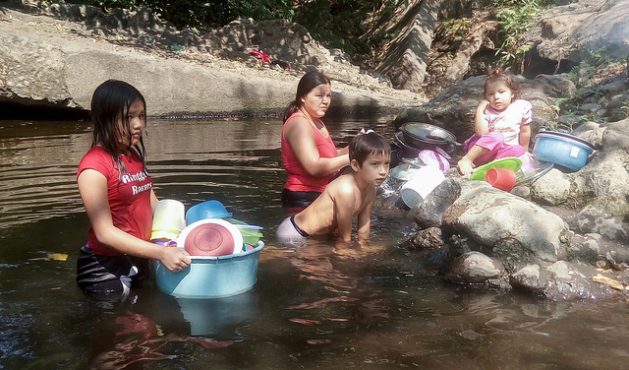 A mother washes kitchen utensils in the Aguas Calientes River, while her children play. She told IPS that this small river, part of the watershed of the Lempa River - the longest in El Salvador - always had an abundant flow, but now due to climate change and the use of water for the irrigation of sugar cane, the water level is down. Credit: Edgardo Ayala/IPS