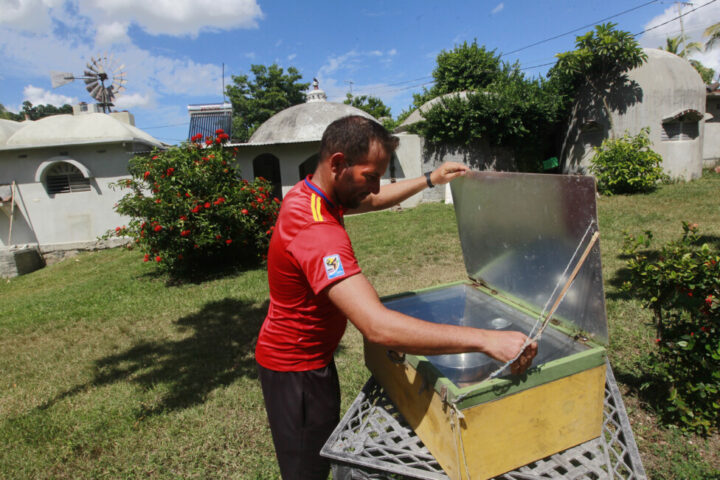 Lorenzo Díaz, the husband of Chavely Casimiro, uses a solar oven to cook food. In the background can be seen a windmill and a solar heater, other technologies that take advantage of the potential for renewable energies on the Finca del Medio farm in central Cuba. CREDIT: Jorge Luis Baños / IPS