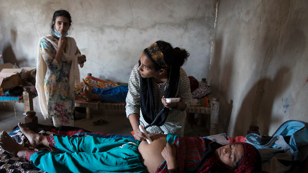 A community health worker examines a pregnant woman inside her home while family members look on, highlighting the growing challenges mothers face as extreme heat and climate-related pressures strain access to healthcare in vulnerable communities. Credit: Wellcome